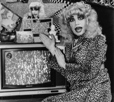 American Music Show character DeAundra Peek sits on set in front of a television and gestures towards it. She is styled in a leopard print dress, heavy makeup, and a blonde wig.