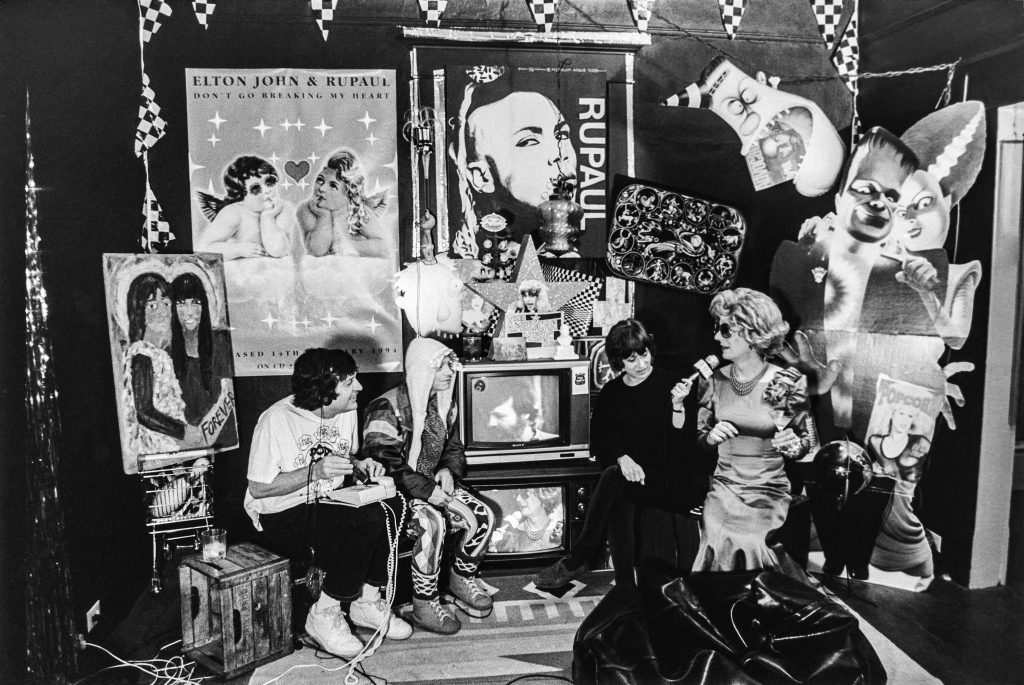 The set of American Music Show with five cast members pictured, including Dick Richards, Duffy Odum, Bud "Beebo" Lowry (on television screen), Potsy Duncan, and Betty Jack Driving. There are paintings, posters, and other ornamentation on the walls and Duncan holds a mic for Driving, who is being interviewed.