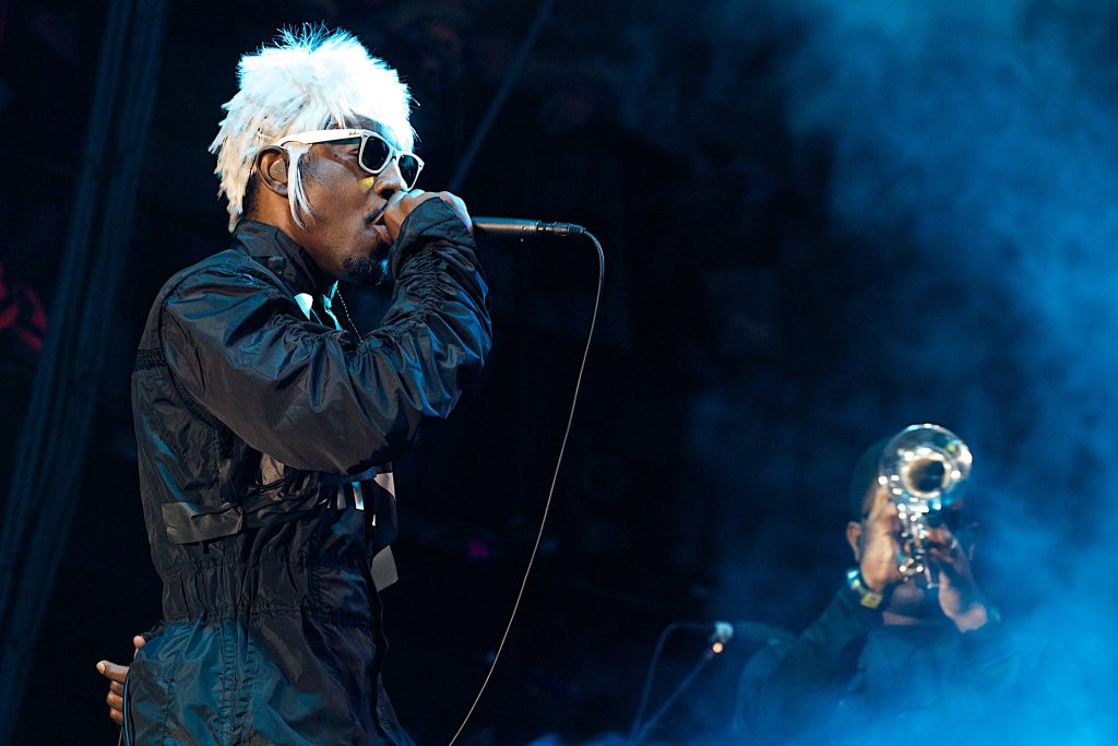 Andre 3000 singing while wearing a white wig and black jumpsuit. A trumpet player stands to the side, shrouded in smoke.