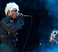 Andre 3000 singing while wearing a white wig and black jumpsuit. A trumpet player stands to the side, shrouded in smoke.